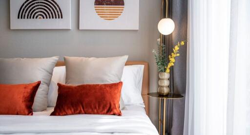 Cozy bedroom with modern decor, featuring a bed with white and orange pillows, a bedside table with a lamp, and artwork on the wall.