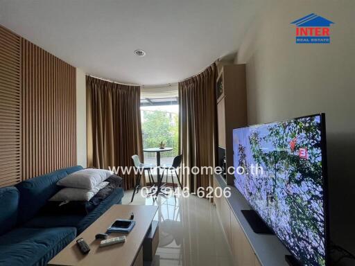 Modern living room with a large blue sofa, flat screen TV, and a dining area by the window