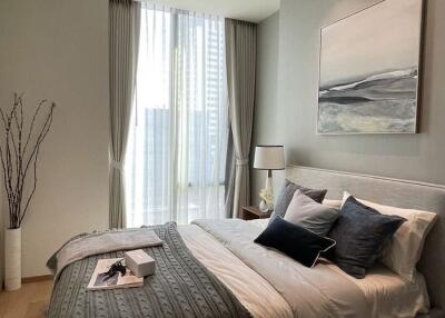 Modern bedroom with natural light, bed with pillows and blanket, bedside table, lamp, and wall art