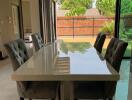 Modern dining room with glass table and upholstered chairs next to large sliding glass doors overlooking a backyard