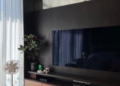 Modern living room with a flat-screen TV on a dark entertainment unit, wooden flooring, and a standing fan with natural light from floor-to-ceiling curtains