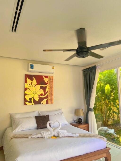 Well-lit bedroom with a bed, air conditioner, ceiling fan, bed lamps, artwork, and curtains