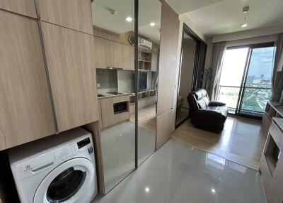 Modern living space with wooden cabinets, washing machine, glass partition, leather sofa, and large window with city view.