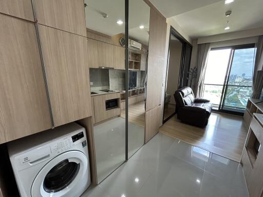 Modern living space with wooden cabinets, washing machine, glass partition, leather sofa, and large window with city view.