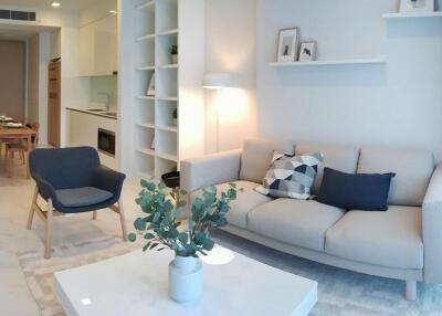 Modern living room with a grey couch, armchair, wall shelves, and a kitchen in the background