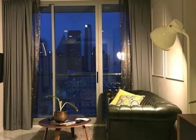 Well-lit living room with large window and evening city view, featuring a green sofa and wall-mounted TV.