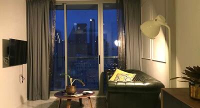 Well-lit living room with large window and evening city view, featuring a green sofa and wall-mounted TV.