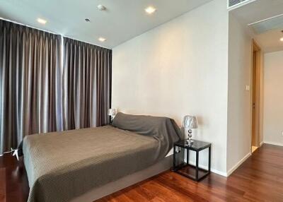 Modern bedroom with wooden floor, bedside tables, lamps, and large windows with dark curtains.