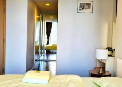 A modern bedroom with a view into the hallway featuring a mirrored closet and a neatly made bed with bedside table and lamp.