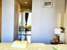 A modern bedroom with a view into the hallway featuring a mirrored closet and a neatly made bed with bedside table and lamp.