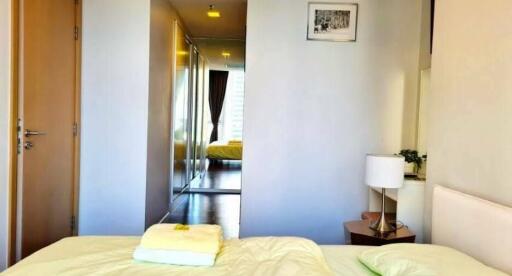 A modern bedroom with a view into the hallway featuring a mirrored closet and a neatly made bed with bedside table and lamp.