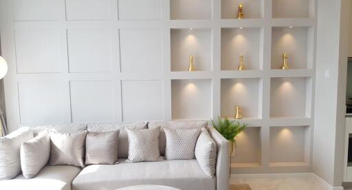 Modern living room with a grey sofa, decorative pillows, built-in shelving with gold ornaments, and a plant