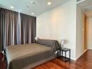 Modern bedroom with hardwood flooring, a bed with brown blanket, side tables with lamps, and dark curtains