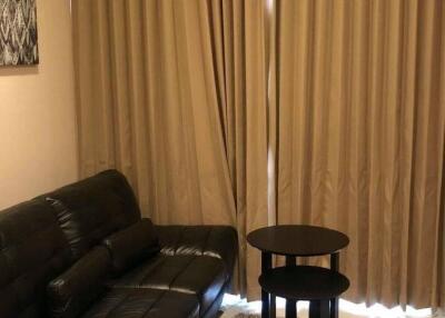 Small living room with a black leather sofa, side tables, wall-mounted TV, and beige curtains