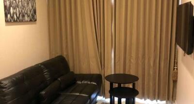 Small living room with a black leather sofa, side tables, wall-mounted TV, and beige curtains