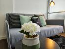 Cozy living room with a sofa and decorative pillows, featuring a coffee table with a flower pot and a floor lamp in the background.