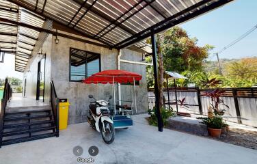 Motorbike parked under a metal roof with a concrete building and a garden in the background