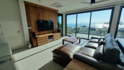 Modern living room with brown leather sofa and wooden TV stand, large windows with ocean view