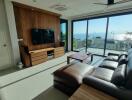 Modern living room with brown leather sofa and wooden TV stand, large windows with ocean view