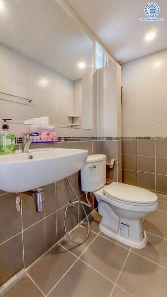 A clean and modern bathroom with gray tiles, a wall-mounted sink, a toilet, and a shelf with toiletries.