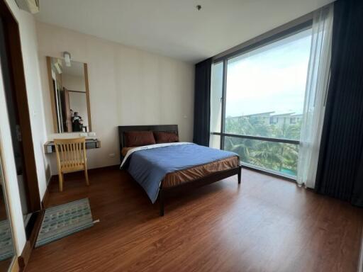 A well-lit bedroom with wooden flooring, a bed, a work desk, and a large window with curtains.