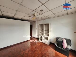 Living room with wooden flooring, white walls, ceiling tiles, a chair, and a small shelving unit.