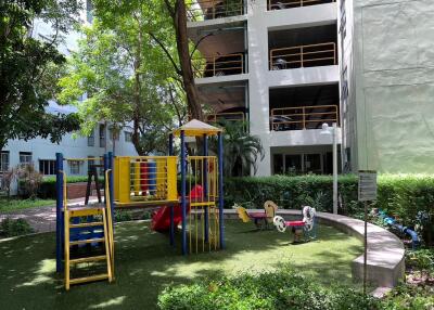Outdoor playground area with slide and seesaws surrounded by greenery and adjacent to multi-story building