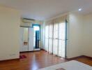 Spacious bedroom with wooden flooring, large windows with sheer curtains, and air conditioning.