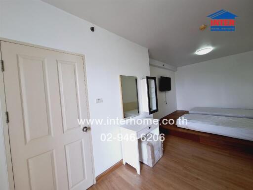 Bedroom with a wooden floor and white walls, featuring a bed, door, TV, mirror, and storage units.