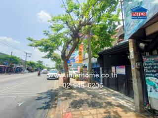 Street view in front of real estate property with a tree, cars, and shops.