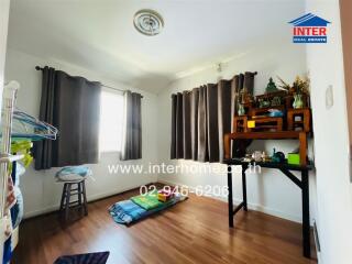 A small bedroom with wooden flooring, brown curtains, and a simplistic setup including a table and a sleeping arrangement on the floor.