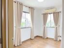 Bedroom with wooden flooring, white walls, and large windows covered by beige curtains