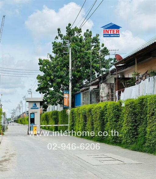 Entrance of residential area with tall green hedge, Inter Real Estate signboard, and road extending into the distance
