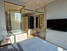 Bright and modern bedroom with a large window providing natural light, a wall-mounted TV, and an adjacent bathroom with a glass partition.