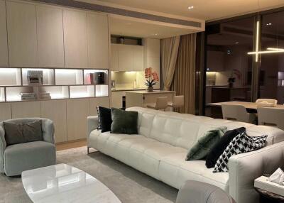 Modern living room with a white sofa, armchairs, a coffee table, dining area, and built-in shelves with lighting