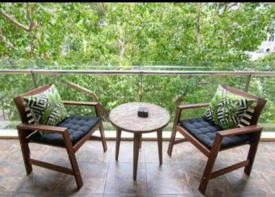 A cozy balcony with two wooden chairs, a small table, and green decorative cushions, overlooking lush trees