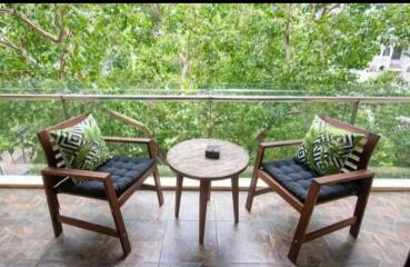 A cozy balcony with two wooden chairs, a small table, and green decorative cushions, overlooking lush trees