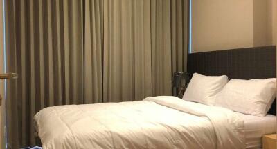 Well-lit bedroom with a large window behind closed curtains, bed with white linens and pillows, and a nightstand.