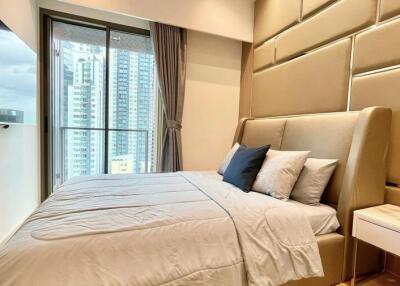 Modern bedroom with a large bed, bedside table, and floor-to-ceiling windows with a city view