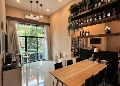 Modern dining area with a wooden table and chairs, shelves with plants and bottles, large window, and cozy ambiance