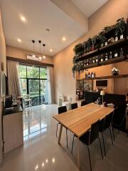 Modern dining area with a wooden table and chairs, shelves with plants and bottles, large window, and cozy ambiance