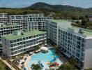 Aerial view of a large residential complex with multiple buildings, green rooftops, and a central swimming pool surrounded by lounge chairs and umbrellas.