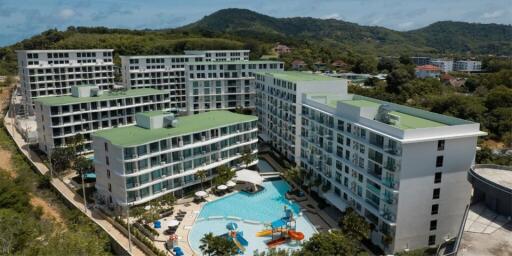 Aerial view of a large residential complex with multiple buildings, green rooftops, and a central swimming pool surrounded by lounge chairs and umbrellas.