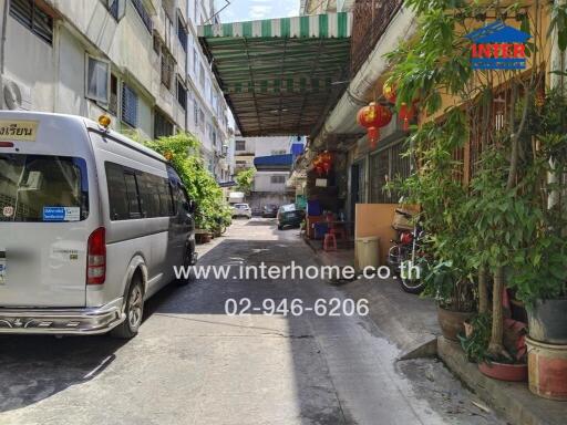 A narrow street with buildings, a parked van, and storefronts with plants