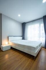 Modern bedroom with wooden flooring, a bed with white linens, bedside table with lamp, and large windows with blue curtains