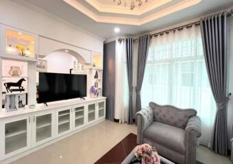 A well-decorated living room with a TV, shelves, and large windows with curtains