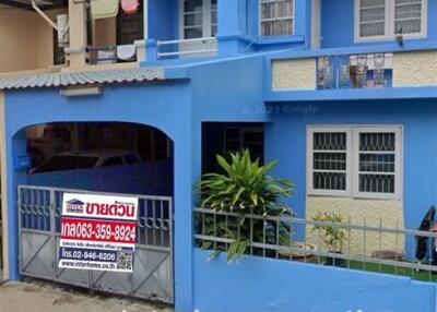 Photo of a two-story house with a blue exterior, red roof tiles, a small front yard, and a fence. The house has a real estate sign in front.