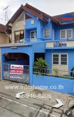 Photo of a two-story house with a blue exterior, red roof tiles, a small front yard, and a fence. The house has a real estate sign in front.