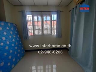 A small bedroom with a large window and a view of neighboring rooftops. The room has light blue curtains, a wall-mounted air conditioner, and a mattress standing against one wall.