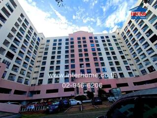 A multi-story residential building with a parking area in front, under a blue sky with clouds.